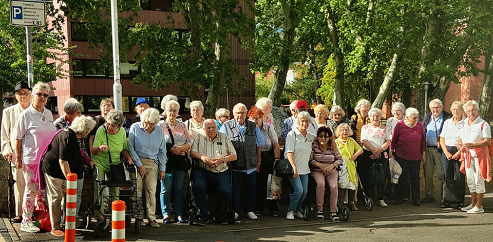 Gruppe älterer Menschen steht an einem sonnigen Straßenrand vor Bäumen und Gebäuden.