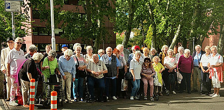 Gruppe älterer Menschen steht an einem sonnigen Straßenrand vor Bäumen und Gebäuden.