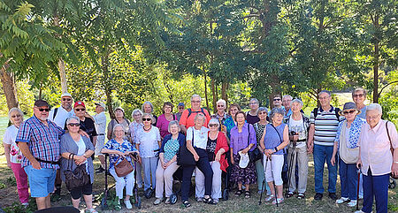 Gruppe von Erwachsenen und Senioren steht im Freien vor Bäumen und Grasfläche bei sonnigem Wetter