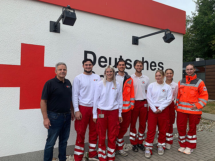 Sieben Personen in einheitlicher Einsatzkleidung vor einer Wand mit großem rotem Kreuz und der Aufschrift "Deutsches Rotes Kreuz"