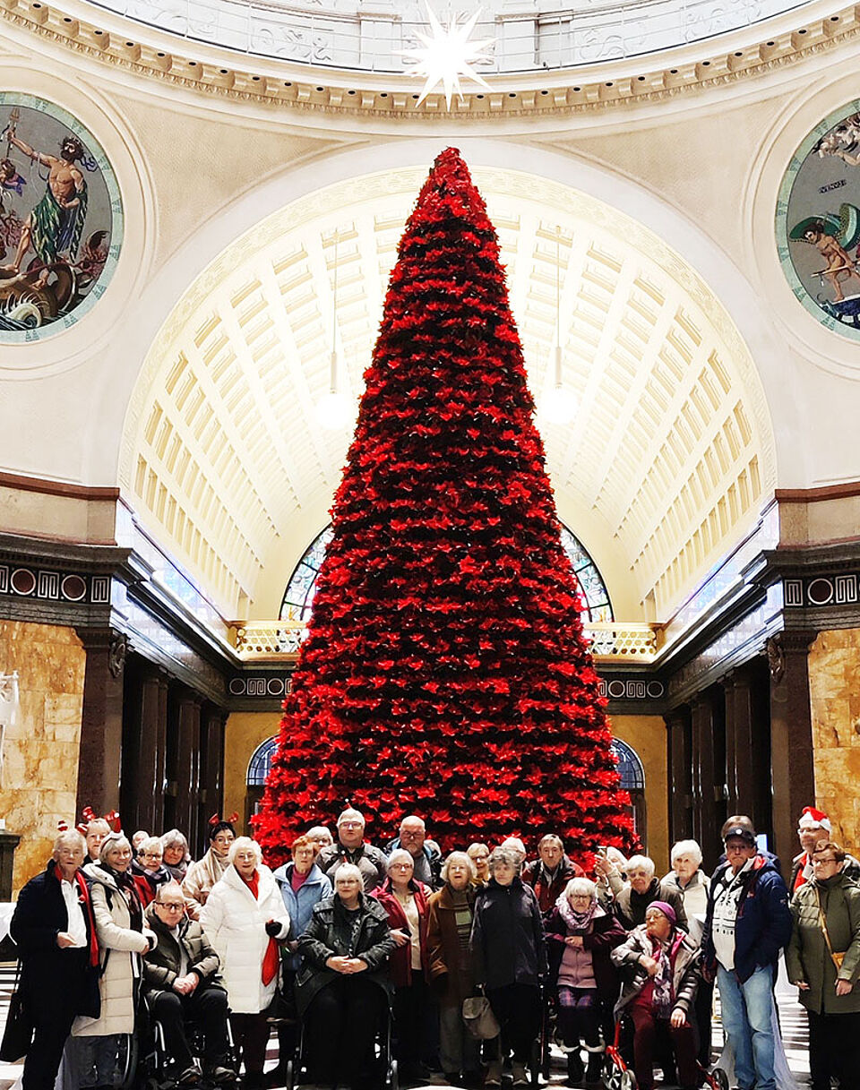 Große Gruppe von Menschen vor einem hohen, rot geschmückten Weihnachtsbaum in einer prunkvollen Halle mit Kuppeldecke.