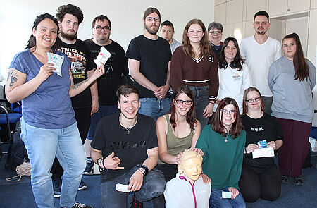 Gruppe von Menschen in einem Raum, einige sitzen auf dem Boden, andere stehen, einige halten kleine weiße Gegenstände in der Hand