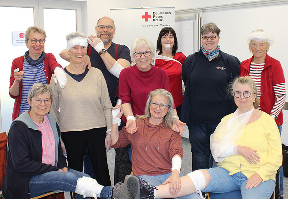 Gruppe von Menschen in einem Raum mit einem Schild des Deutschen Roten Kreuzes im Hintergrund, einige Personen haben Verbände an Armen oder Beinen