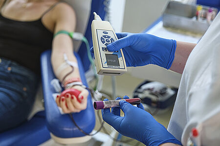 Person sitzt mit ausgestrecktem Arm und rotem Stressball während einer Blutspende, daneben hält eine Person mit blauen Handschuhen ein Messgerät und ein mit Blut gefülltes Röhrchen.