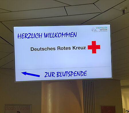 Leuchtschild mit dem Text 'HERZLICH WILLKOMMEN Deutsches Rotes Kreuz' und einem roten Kreuz sowie einem blauen Pfeil und dem Text 'ZUR BLUTSPENDE'
