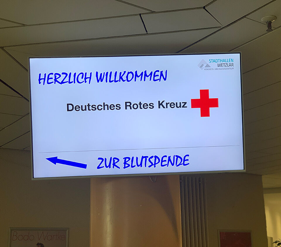 Leuchtschild mit dem Text 'HERZLICH WILLKOMMEN Deutsches Rotes Kreuz' und einem roten Kreuz sowie einem blauen Pfeil und dem Text 'ZUR BLUTSPENDE'