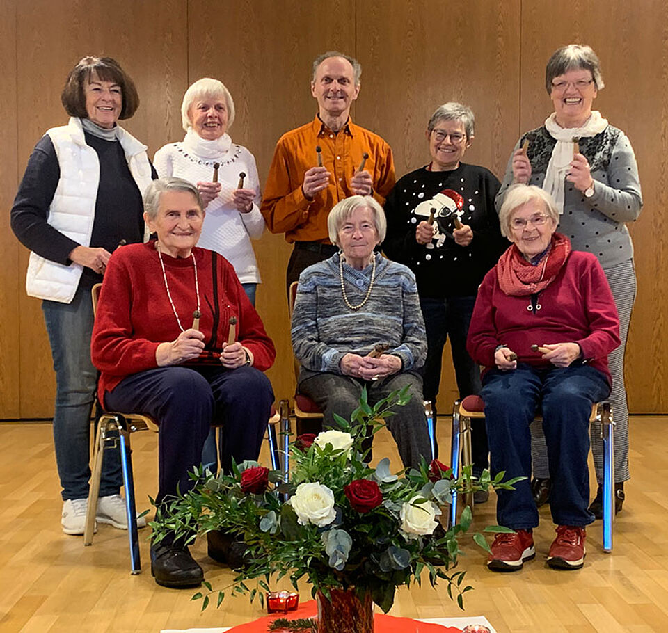 Gruppe von sieben Personen, drei sitzen auf Stühlen, vier stehen dahinter, alle halten kleine Glöckchen, davor ein Blumenarrangement auf dem Boden.