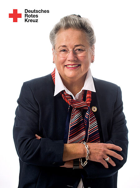 Brigitte Wettengel Person mit verschränkten Armen trägt dunkles Sakko, weißes Hemd und gestreiften Schal vor weißem Hintergrund mit Logo des Deutschen Roten Kreuzes