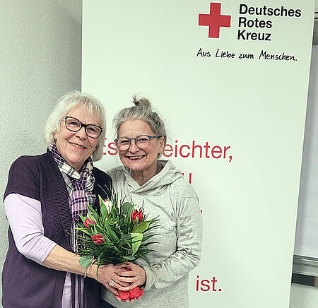 Zwei Frauen stehen vor einem Banner mit dem Logo des Deutschen Roten Kreuzes und halten gemeinsam einen Blumenstrauß